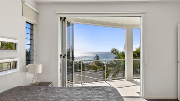 A bedroom with a bed, a nightstand, a lamp, and a sliding glass door leading to a balcony with a view of the ocean. - Relax in stylish comfort, Sunshine Beach (Sunshine Beach)