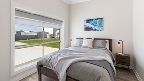 A bedroom with a large window, a bed with a grey blanket, a bedside table with a lamp, and a wall-mounted artwork.