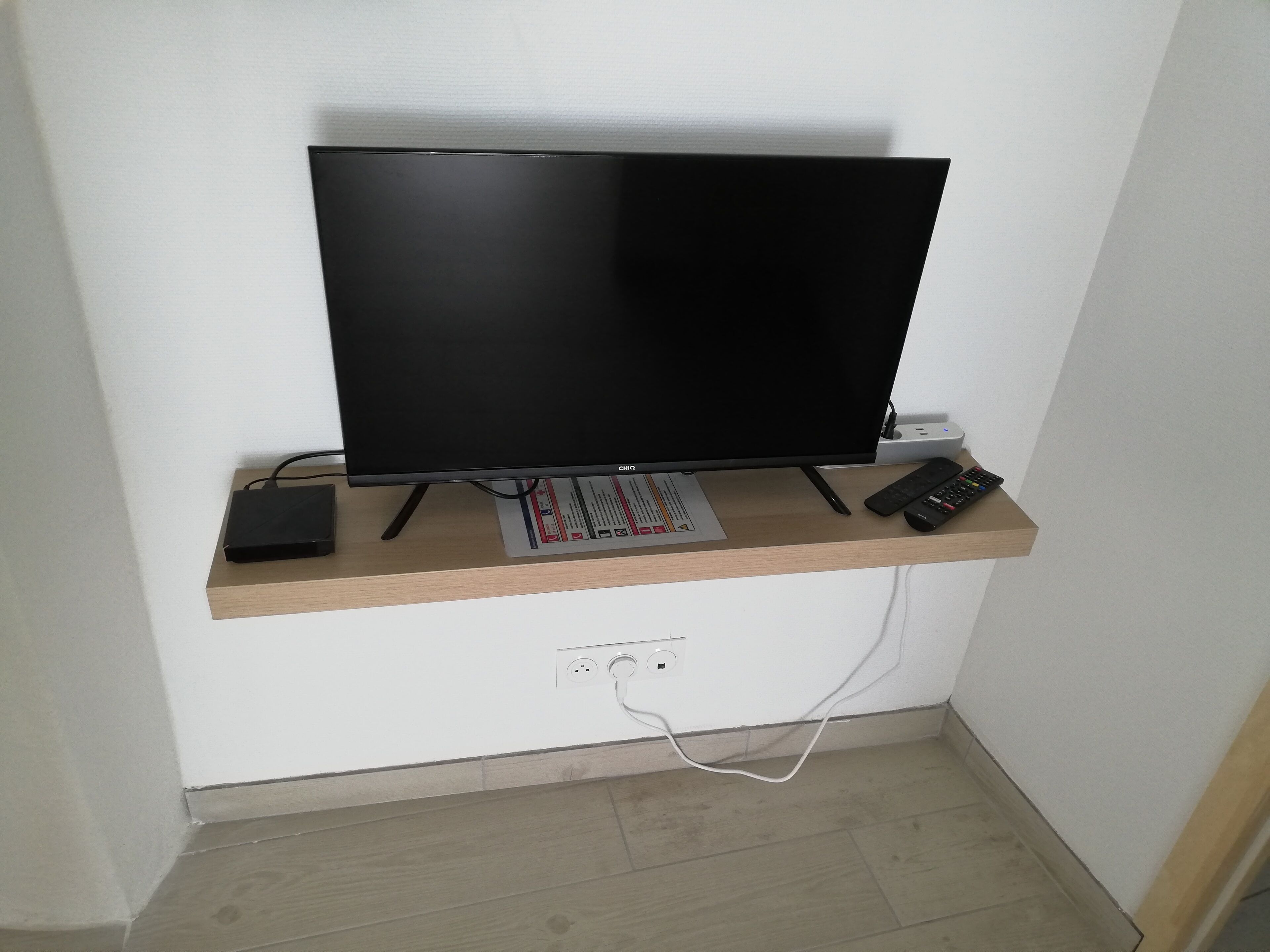Ein Flachbildfernseher auf einem Holzregal, darunter eine Fernbedienung und eine Steckschienenleiste.