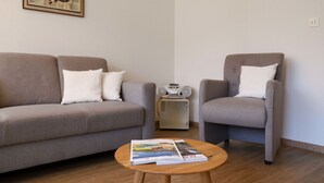 Un salon avec un canapé gris, deux fauteuils, une table basse avec des magazines, une radio et deux tableaux accrochés aux murs.
