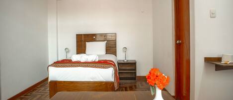 Quarto de hotel com cama de madeira, mesa de cabeceira, luminária e um vaso com flores, além de piso de madeira.