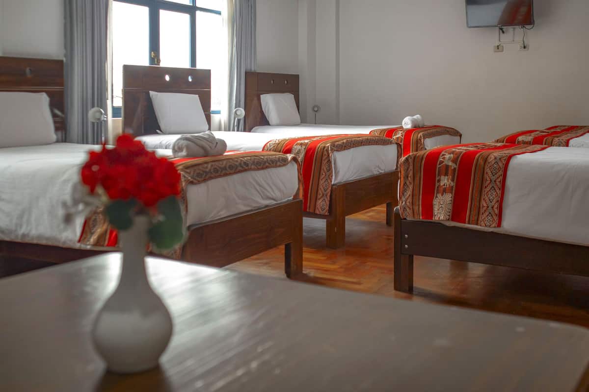 A hotel room with three beds, each with a wooden headboard and white bedding. There is a vase with red flowers on a wooden table in the foreground.