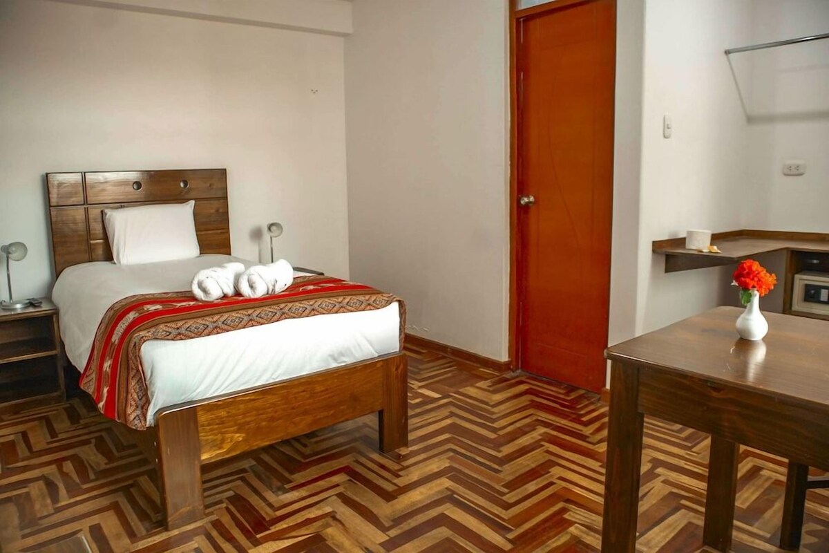 Habitación de hotel con cama de madera, mesita de noche, una silla, suelo de madera y una puerta.