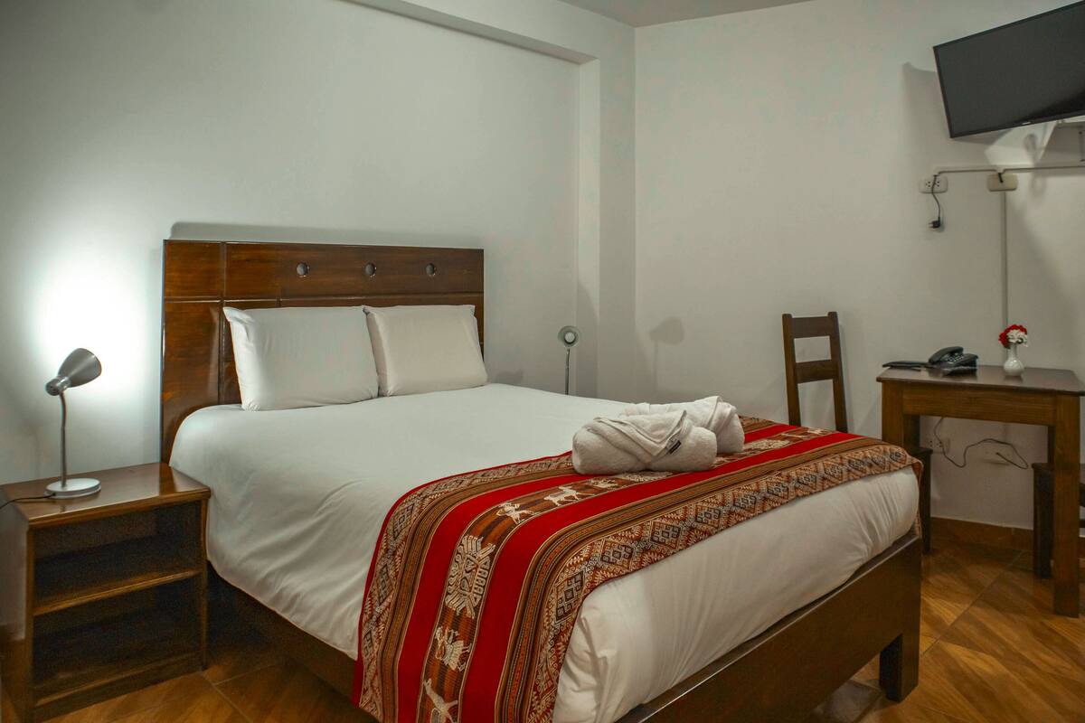 A neatly made bed with a patterned blanket, a bedside lamp, a wooden nightstand, a desk with a chair, and a flat-screen TV mounted on the wall.