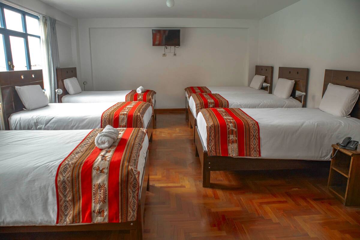 A hotel room with four single beds, each with a red and white patterned bedspread, a white pillow, and a rolled towel on top. There is a wooden floor, a small wooden nightstand with a phone, and a wall-mounted TV.