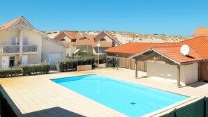 A swimming pool surrounded by houses with red-tiled roofs, a clear blue sky, and a hilly landscape in the background.