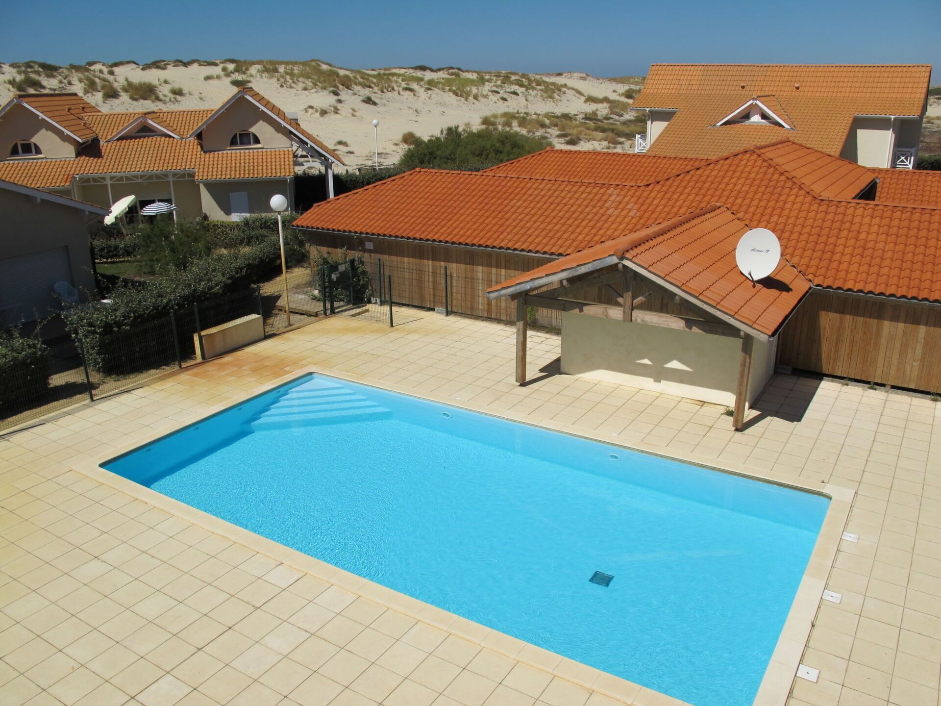 Une piscine avec une terrasse carrelée, entourée de maisons aux toits recouverts de tuiles rouges, et un paysage sablonneux en arrière-plan.
