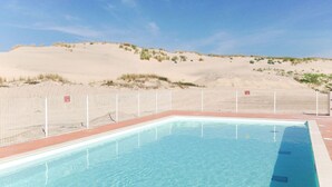 A rectangular swimming pool with clear blue water, surrounded by a white fence, set against a backdrop of sand dunes and sparse vegetation under a clear sky. - Cosy apartment for 4 guests with WIFI, pool, TV, terrace and pets allowed (Biscarrosse)