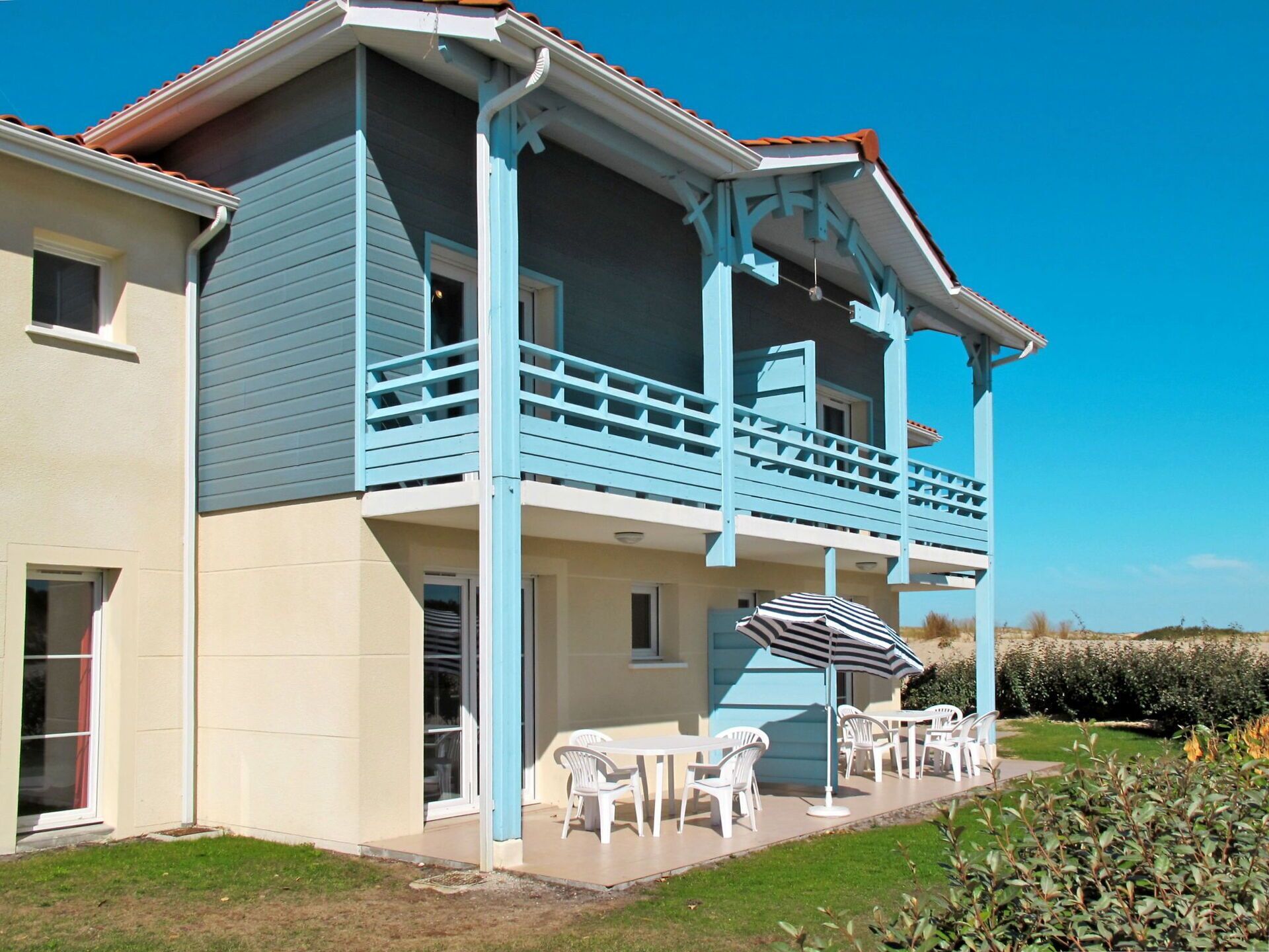 Een tweelaags huis met een balkon en een terras met tuinmeubilair, onder een helderblauwe lucht.