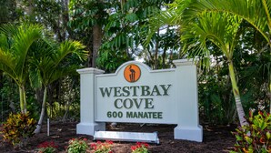 A sign for Westbay Cove, 600 Manatee, surrounded by lush greenery and flowering plants.