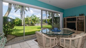 A room with a glass table, wicker chairs, a TV, and a view of a green lawn and palm trees.