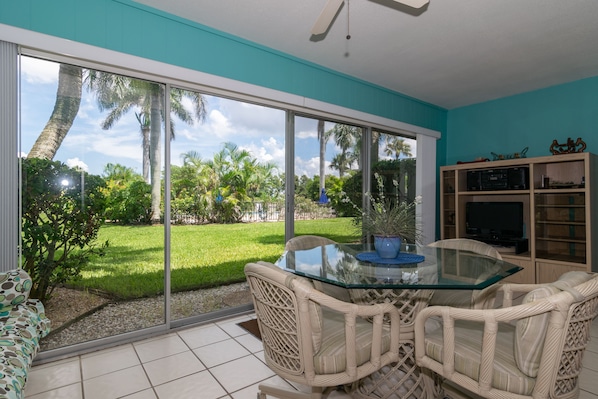 A room with a glass table, wicker chairs, a TV, and a view of a green lawn and palm trees.