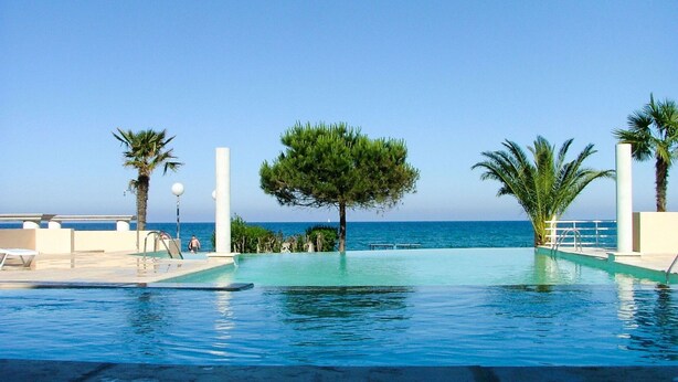 An infinity pool with a clear view of the sea, surrounded by palm trees and a white railing. - Nice apartment for 6 people with A/C, pool, internet, TV, balcony and pets allowed (Santa-lucia-di-moriani)