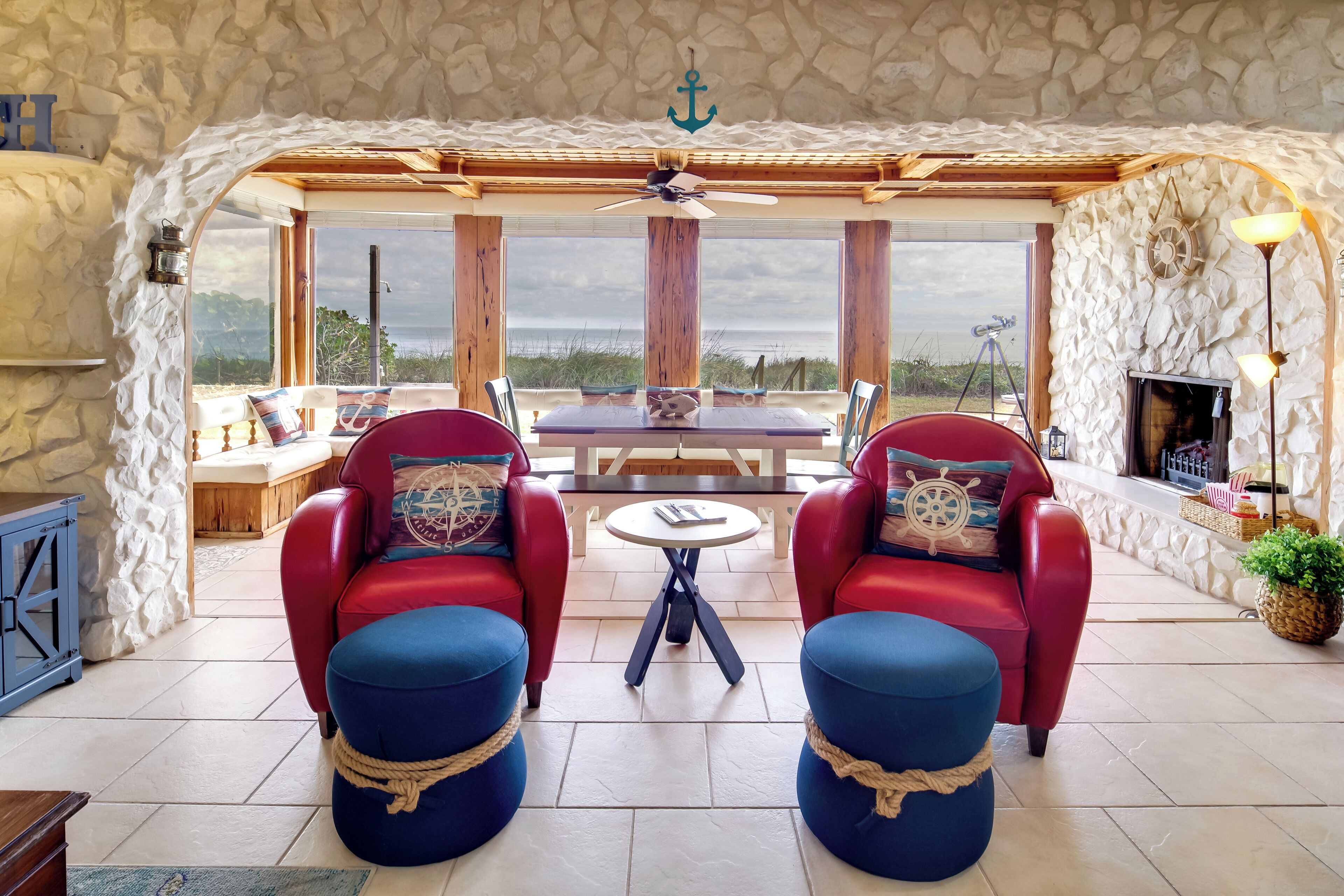 A cozy seating area with red chairs and blue ottomans, a wooden table, and a stone wall with a view of the beach.