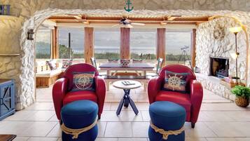Ein gemütlicher Sitzbereich mit roten Stühlen und blauen Hocker, einem Holztisch und einer Steinwand mit Blick auf den Strand.