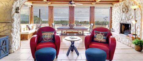A cozy seating area with red chairs and blue ottomans, a wooden table, and a stone wall with a view of the beach.