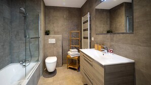 Een moderne badkamer met een douchecabine van glas, een wit toilet, een houten krukje en een wastafel met spiegel.