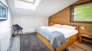 Een slaapkamer met een houten hoofdeinde, een bed met witte lakens, een grijze stoel, een klein nachtkastje met een lamp, en uitzicht op groen door een raam.