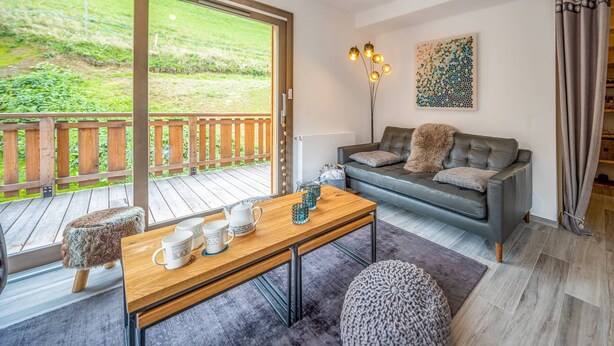 Een gezellige woonkamer met een houten terras, een grijze bank met een bont plaid, een bijzettafeltje met mokken en een schilderij aan de muur.