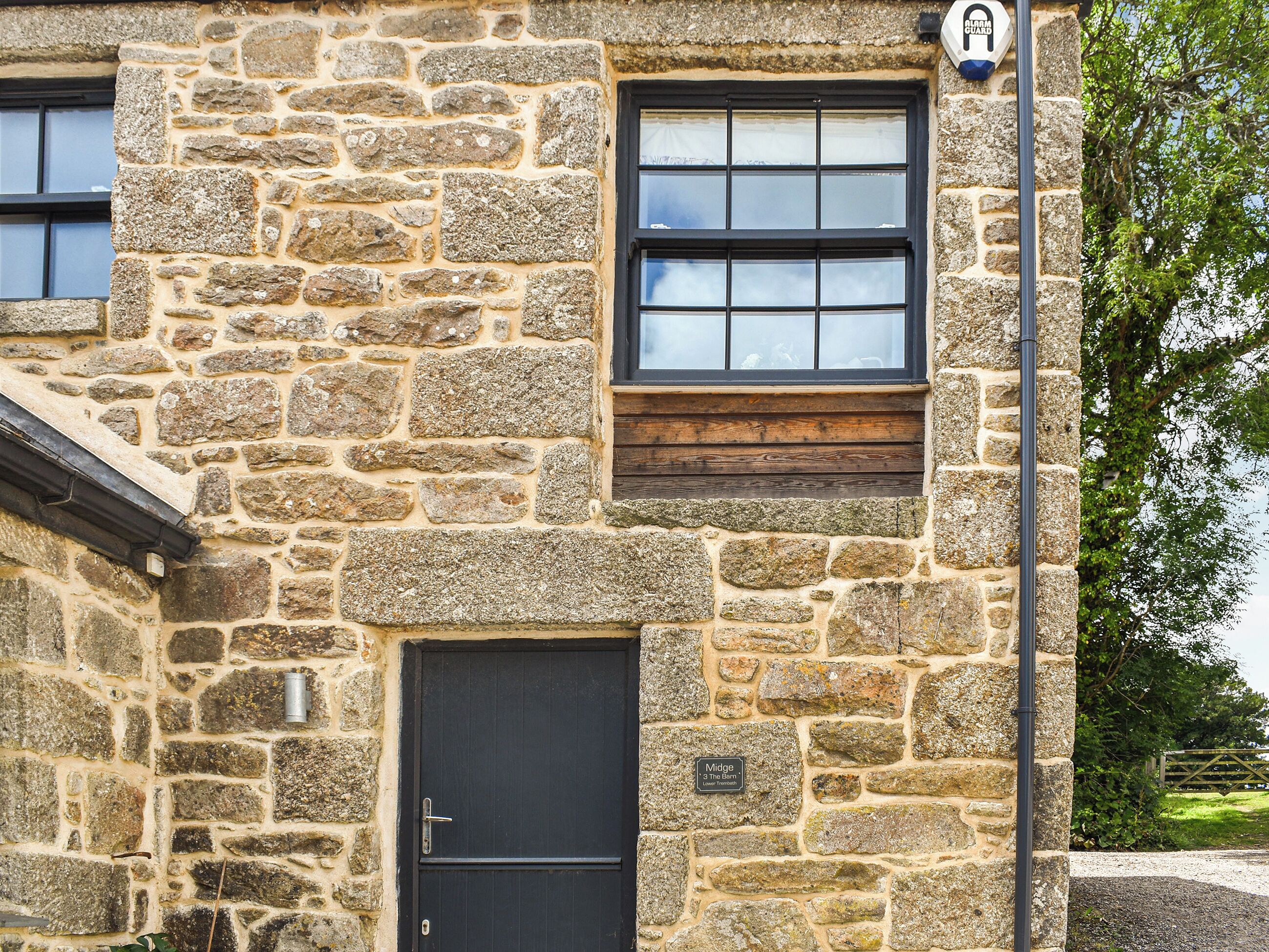 A stone building with a black door and windows, a sign indicating 'Midge 3 The Barn,' and a 'Gratia Guard' sign on a pole.