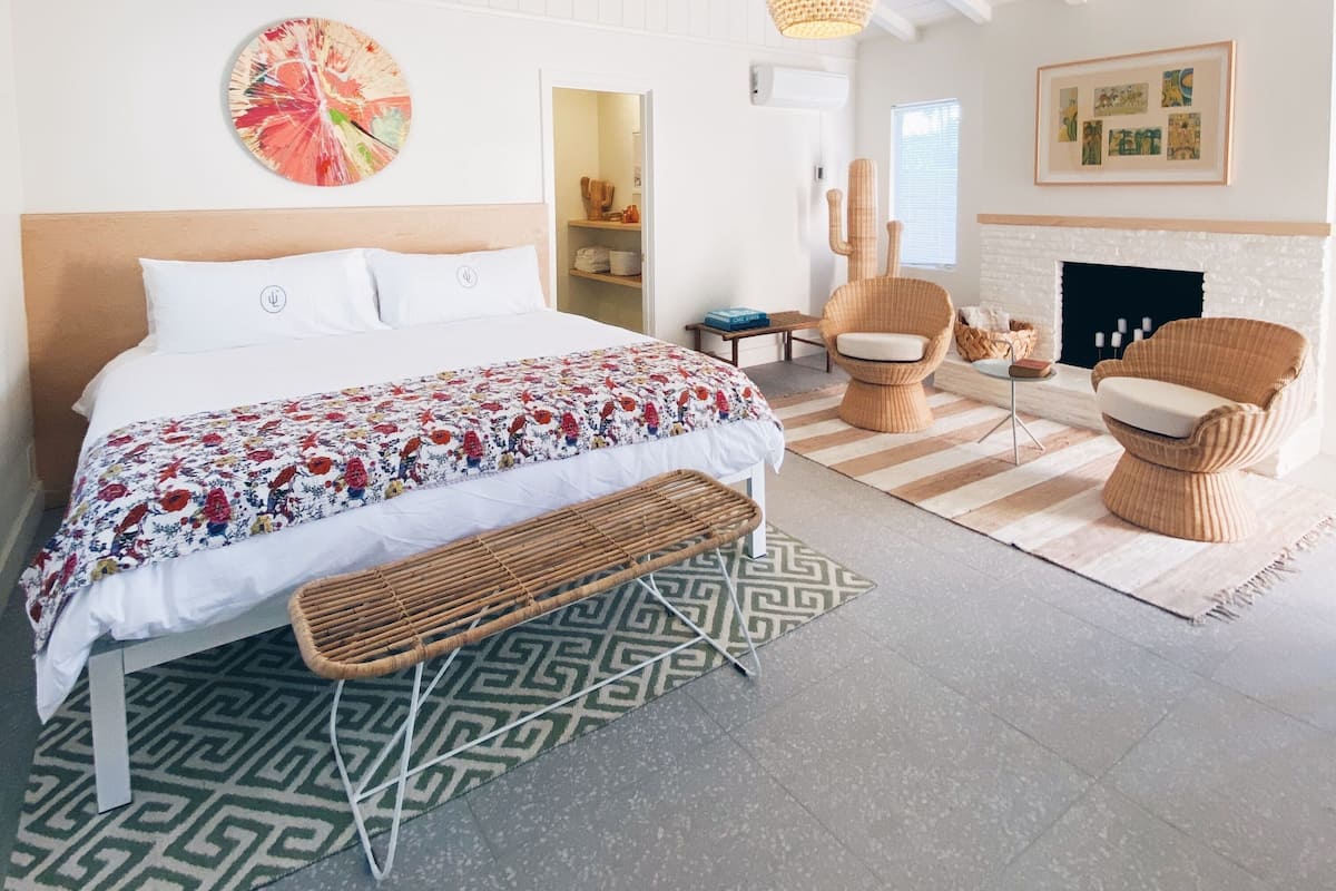 A spacious bedroom with a large bed, a wicker bench, a fireplace, and a ceiling with exposed beams.