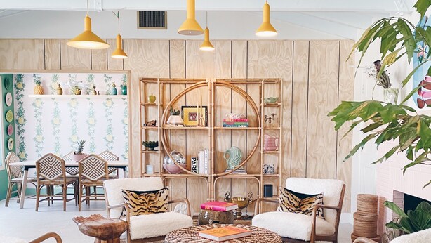 A cozy living room with a round woven ottoman, wooden chairs, a bookshelf, and hanging yellow pendant lights. - Les Cactus (Palm Springs)
