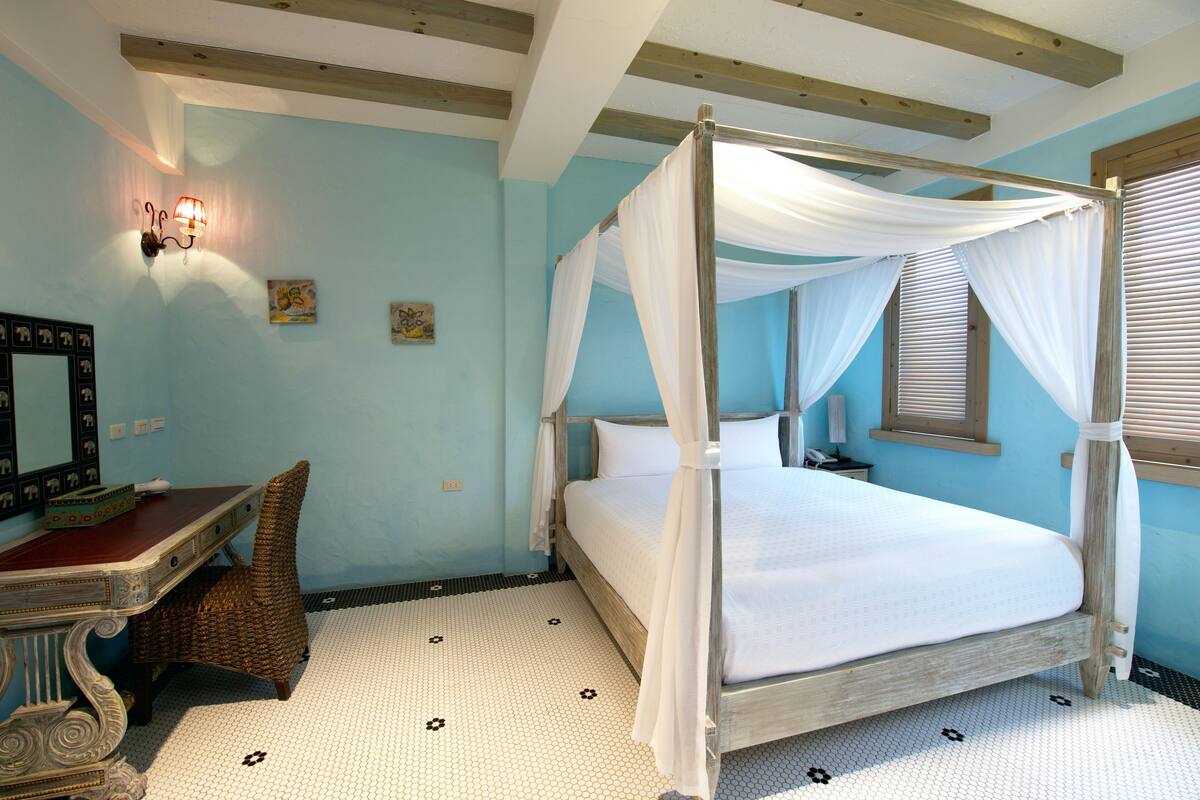A four-poster bed with white linens, a wooden desk with a mirror, and a wicker chair.