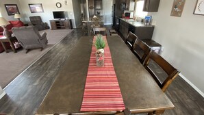 A dining area with a long table, chairs, and a red-striped runner. There is a kitchen in the background with dark cabinets and a window. -  50% to Book the Gecko Home - Super Clean, Really Nice 3BR - Sleeps 6- By TNT. (Page)