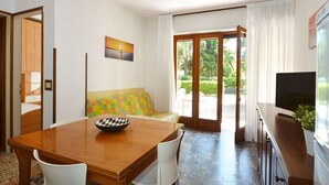 A room with a wooden dining table, white chairs, a colorful sofa, a bowl on the table, a TV on a cabinet, and a sliding glass door leading to an outdoor area. - Nice apartment for 6 people with A/C, TV, terrace and pets allowed (San Michele Al Tagliamento)