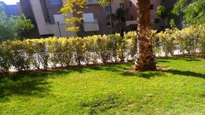 A well-maintained grassy area with a palm tree and neatly trimmed bushes, surrounded by a building with multiple balconies. - Apartment Marrakech Prestigia Golf City 4601 (Marrakech)