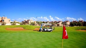 A golf course with a flag, a golf cart, and buildings in the background. - Apartment Marrakech Prestigia Golf City 4601 (Marrakech)