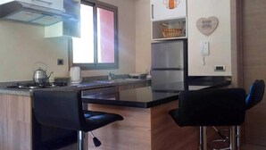 A modern kitchen with a gas stove, a kettle, and a refrigerator. There are two black bar stools and a window providing natural light. - Apartment Marrakech Prestigia Golf City 4601 (Marrakech)