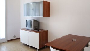 Una habitación pequeña y limpia con una mesa de madera, sillas blancas, un televisor en un mueble y una ventana con cortinas.