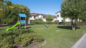 Un parque infantil con un tobogán y estructuras para escalar, rodeado de césped y árboles cuidados, con edificios residenciales al fondo.