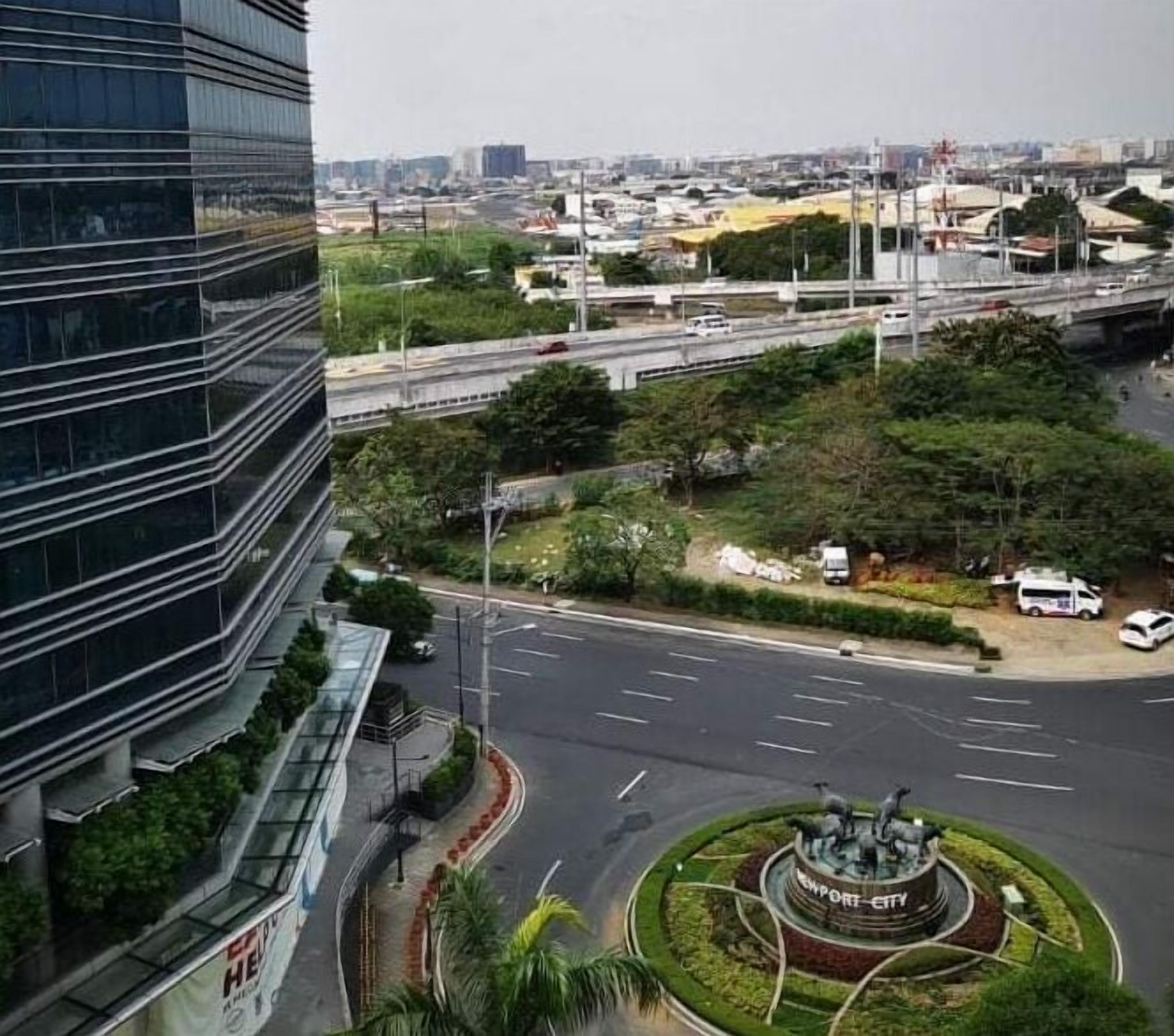 A modern urban intersection with a central roundabout, a statue, and a multi-level highway above.