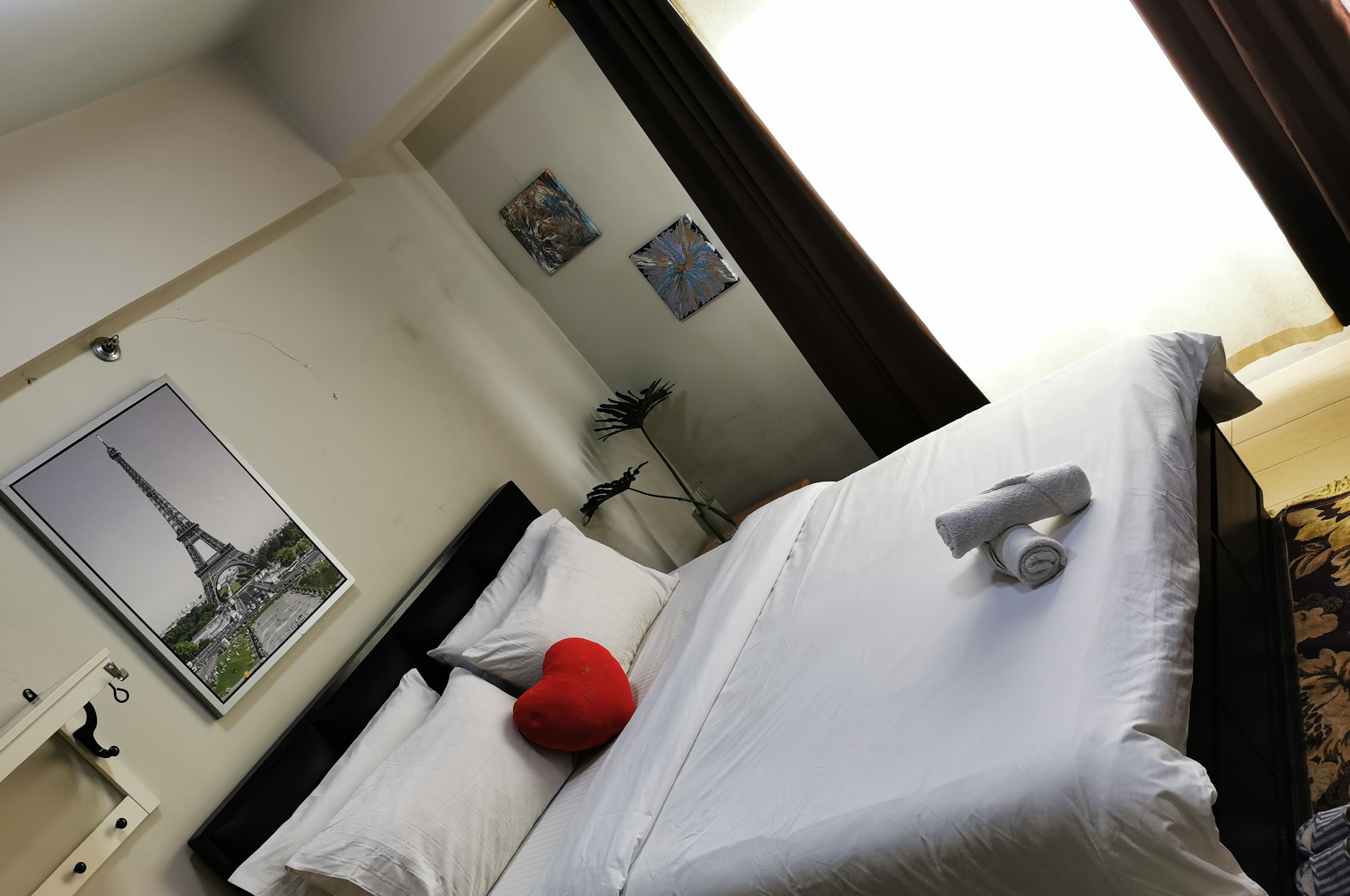 A hotel room with a bed, white linens, a red pillow, a rolled towel, a picture of the Eiffel Tower, and a small plant.