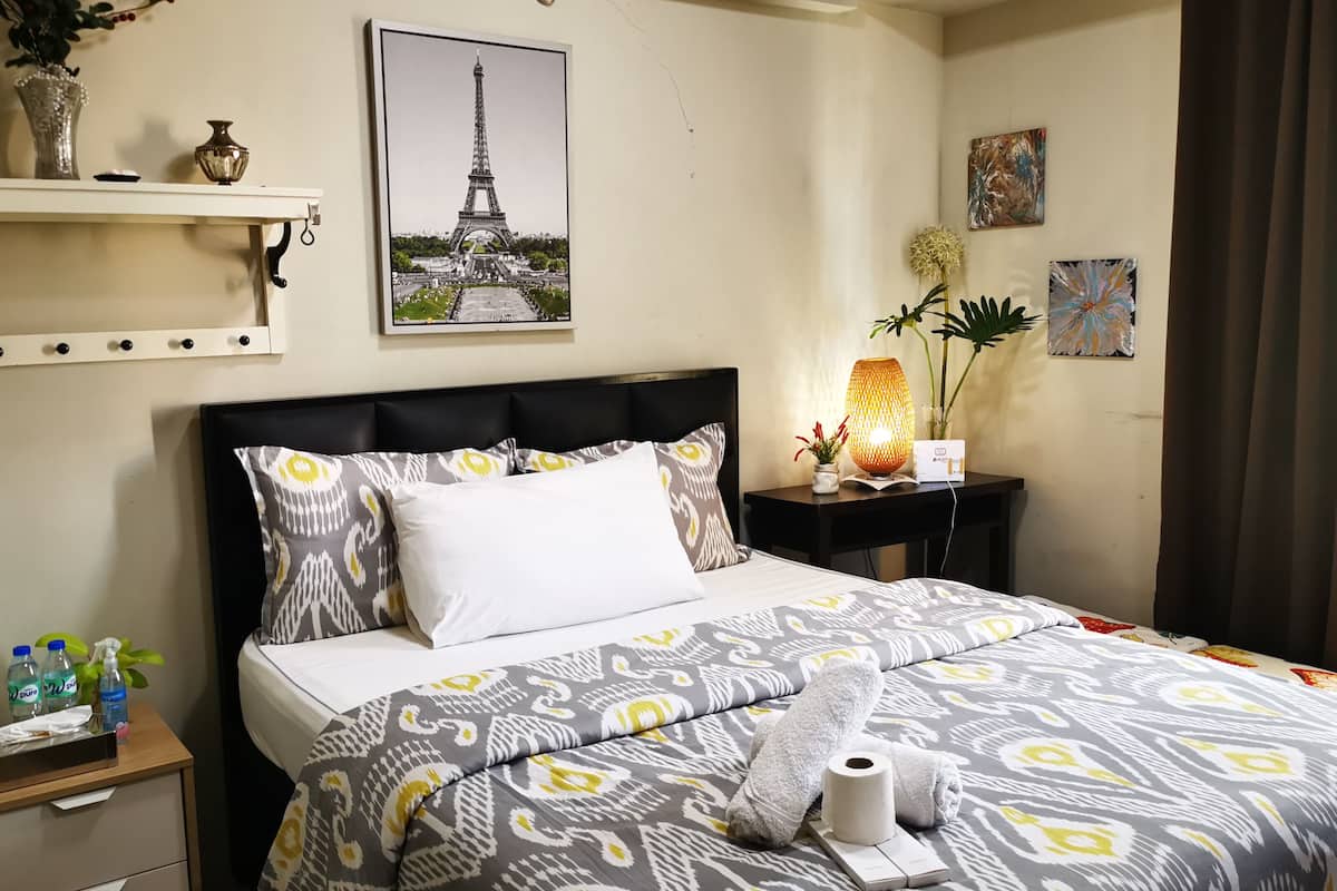 Una cama bien hecha con ropa de cama estampada, una mesita de noche con artículos de aseo, un estante en la pared con adornos, y un cuadro con la Torre Eiffel.