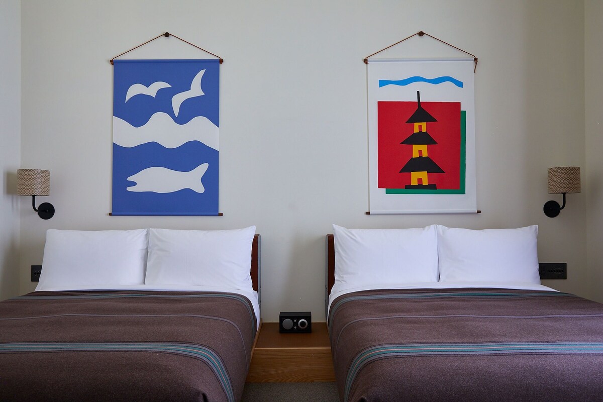 Dos camas con almohadas blancas y ropa de cama marrón, un radio montado en la pared y dos obras de arte en la pared.
