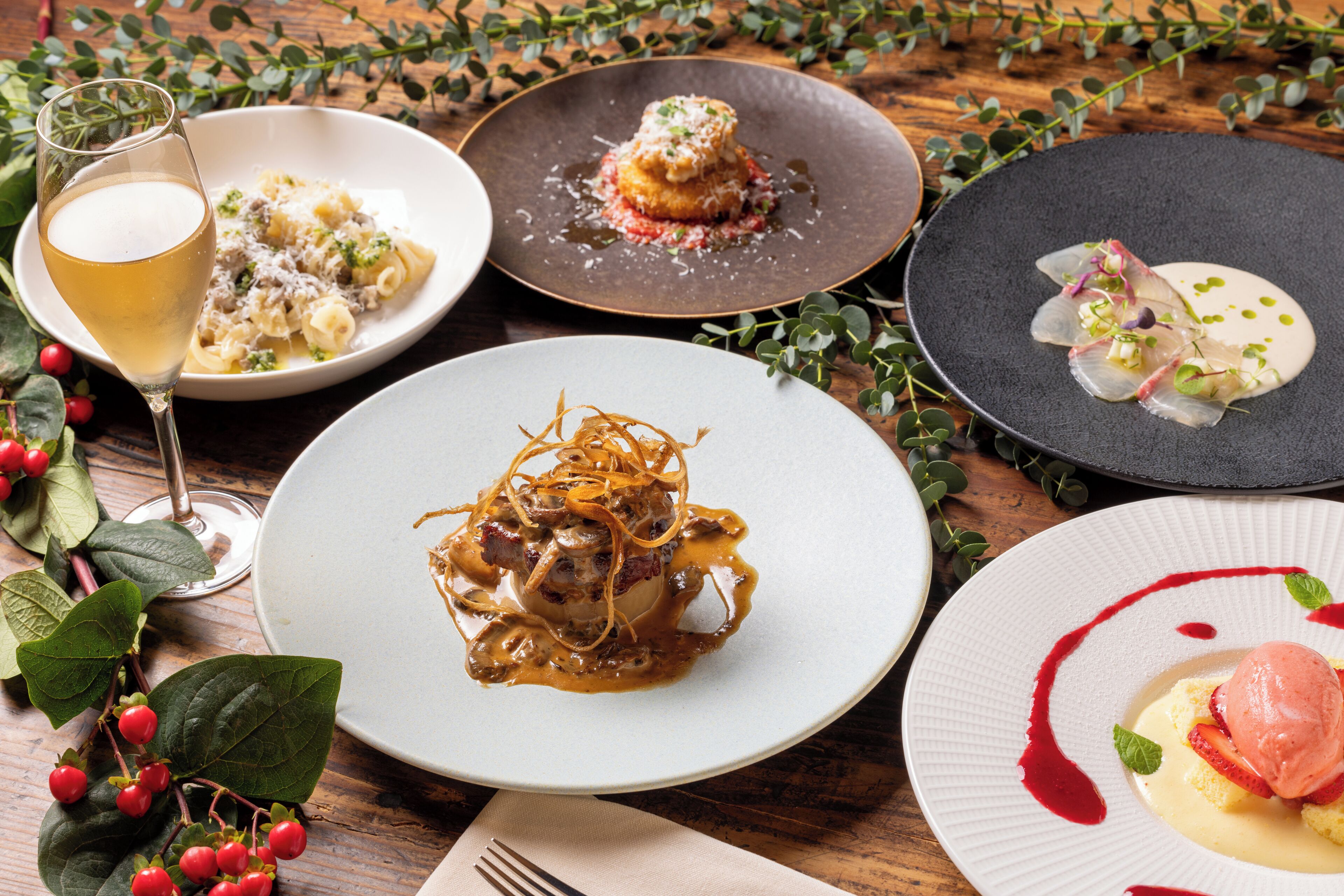 Une variété de plats présentés sur des assiettes, un verre de vin et quelques éléments de verdure sur une table en bois.