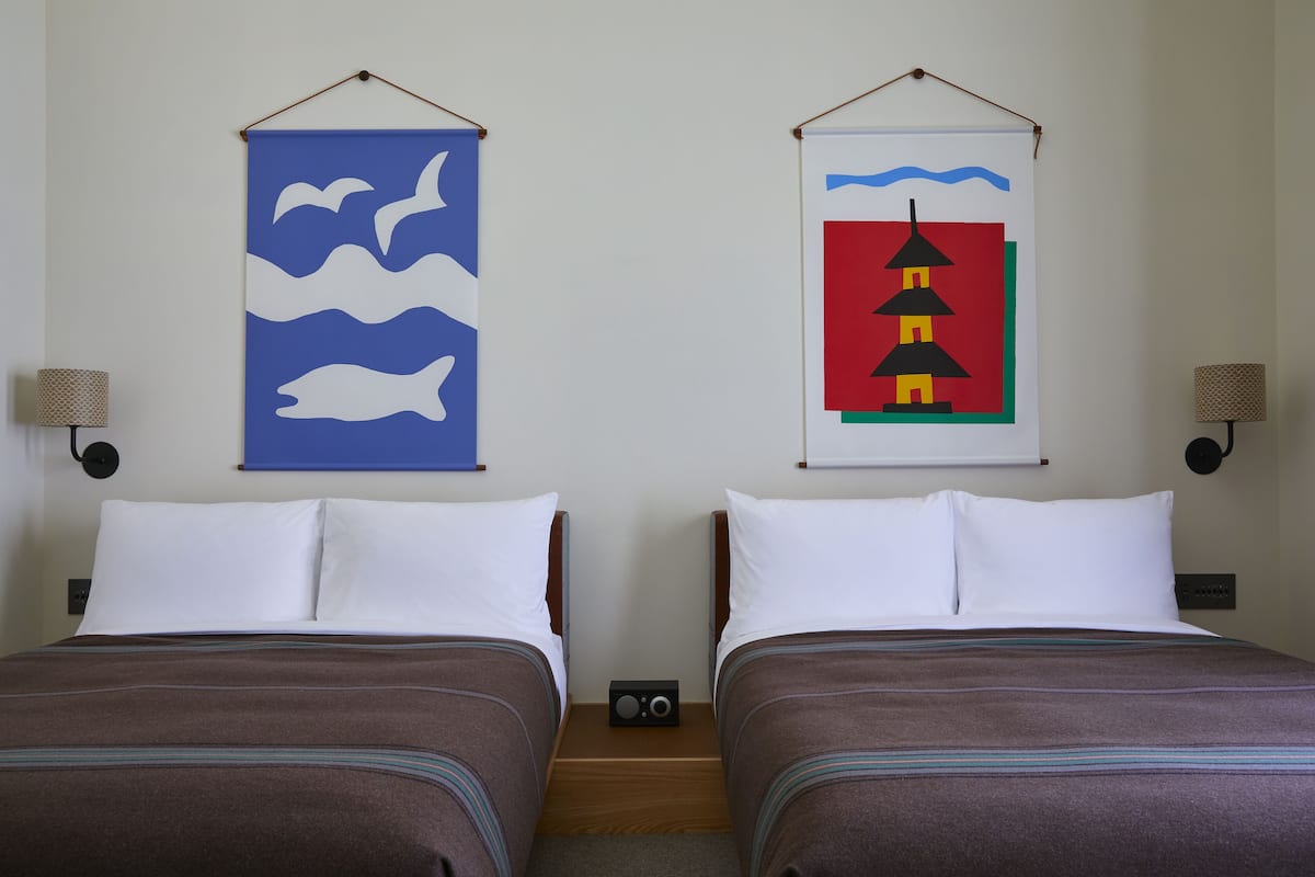 Une chambre d’hôtel avec deux lits, chacun agrémenté d’oreillers blancs et de draps bruns. Deux lampes murales sont fixées de chaque côté des lits.