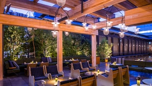 Un espace extérieur aménagé avec des tables et des chaises en bois, entouré de grandes fenêtres et surmonté d’une structure de plafond en bois.