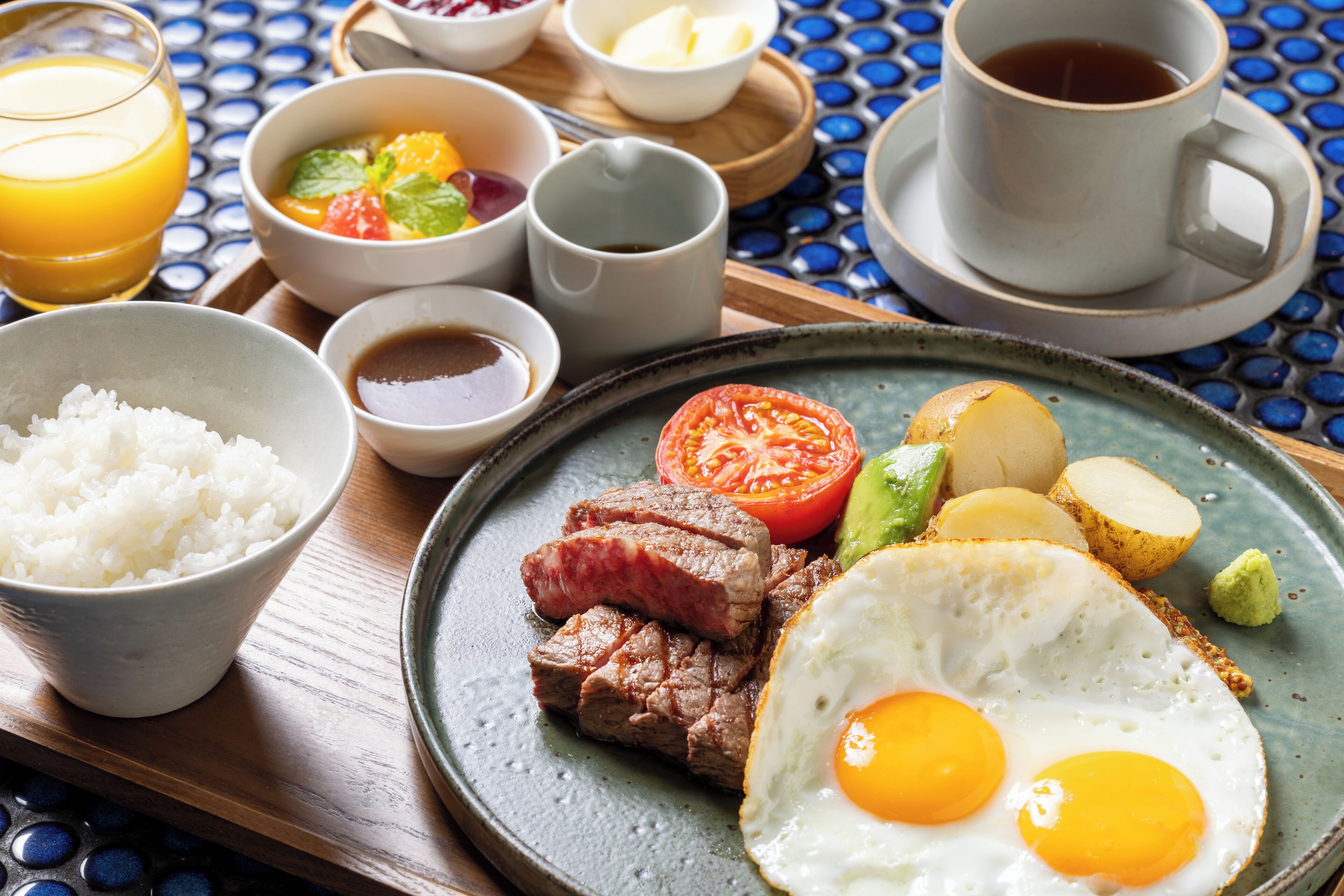 A breakfast plate with steak, eggs, and vegetables, accompanied by bowls of rice and soup, and a cup of coffee on a table with a blue patterned tablecloth.