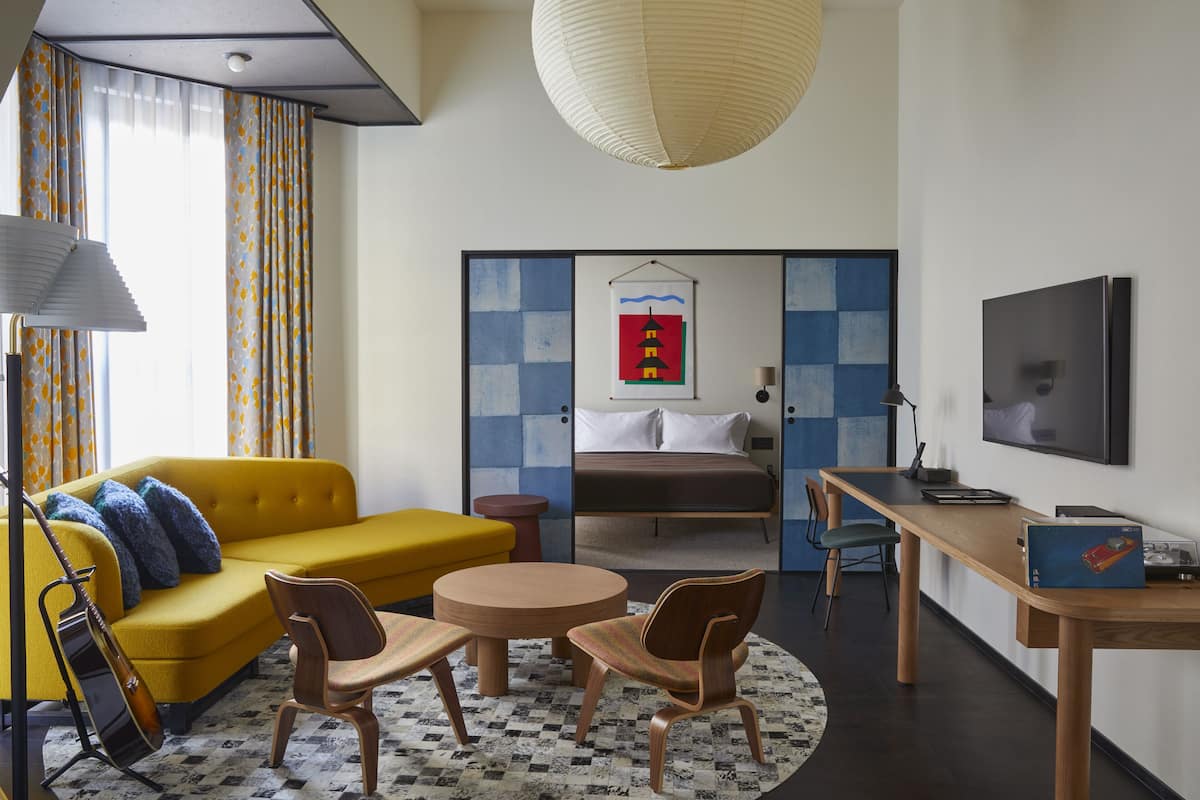 Une chambre d’hôtel moderne avec un canapé jaune, une table à manger en bois, un lit doté d’une tête de lit à carreaux bleus et blancs, et un téléviseur fixé au mur.