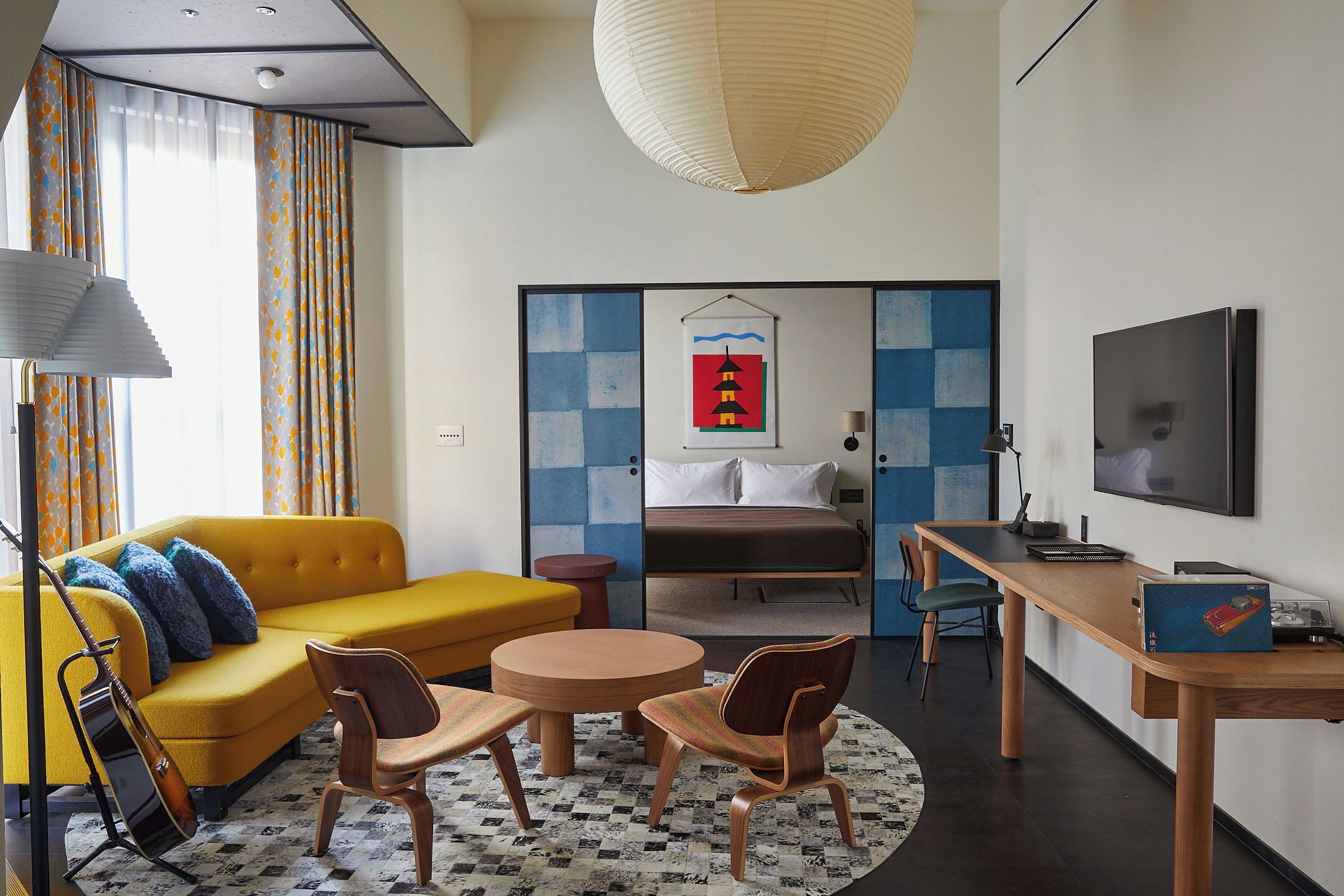 A modern hotel room with a sofa, chairs, a desk, a bed, a TV, and artwork on the walls.