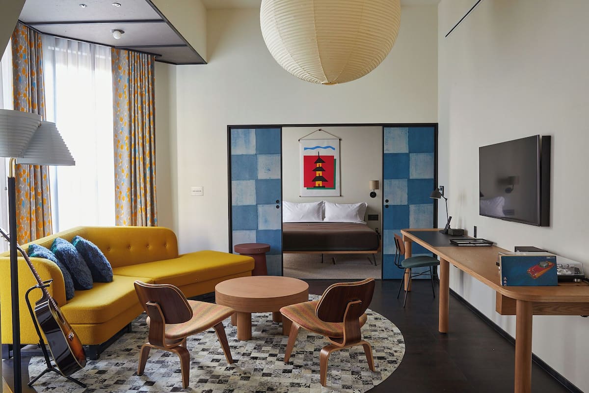 Une chambre d’hôtel moderne, équipée d’un canapé, de fauteuils, d’un bureau, d’un lit, d’une télévision et d’œuvres d’art accrochées aux murs.