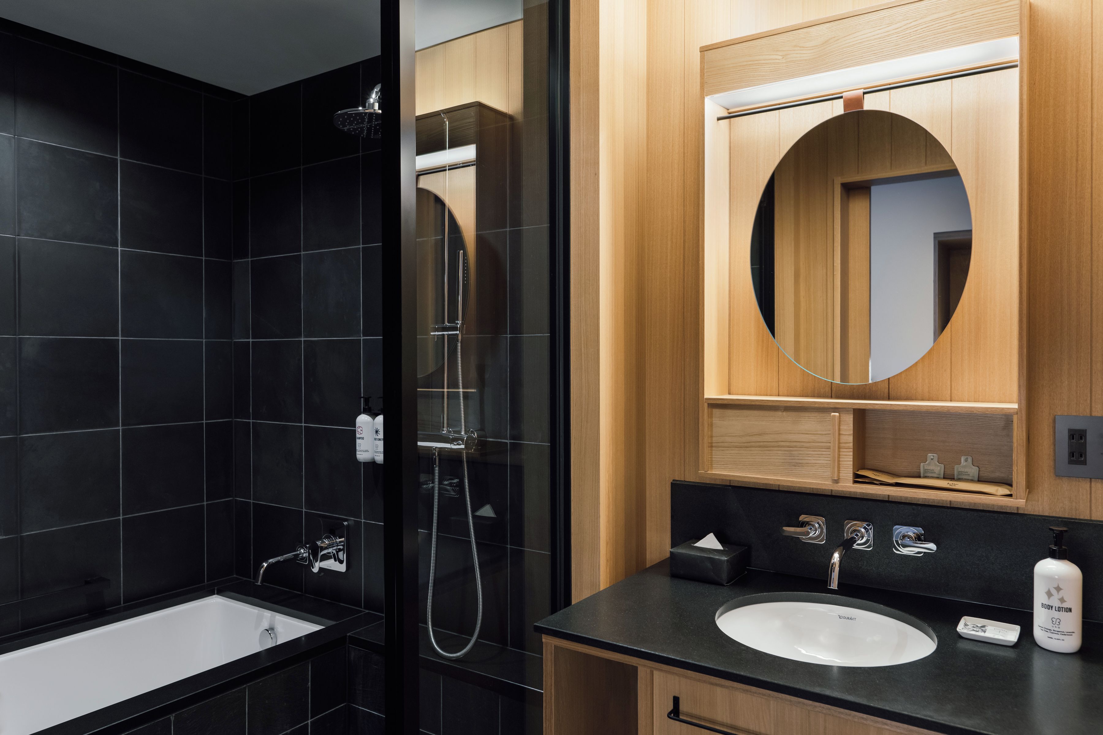 A modern bathroom with a black tiled shower, a white sink, and a wooden vanity.