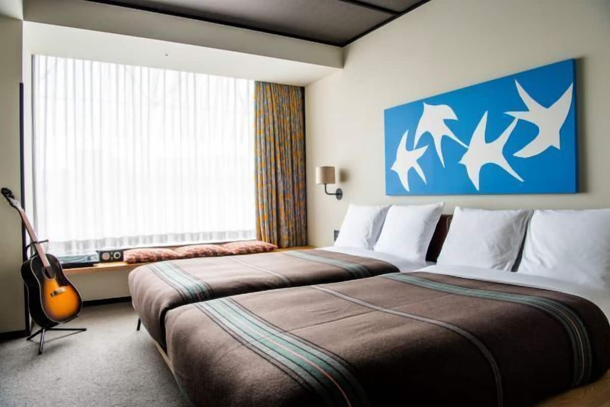 Une chambre d’hôtel avec deux lits, une guitare, une fenêtre avec des rideaux et un mur bleu orné d’une œuvre d’art abstraite blanche.