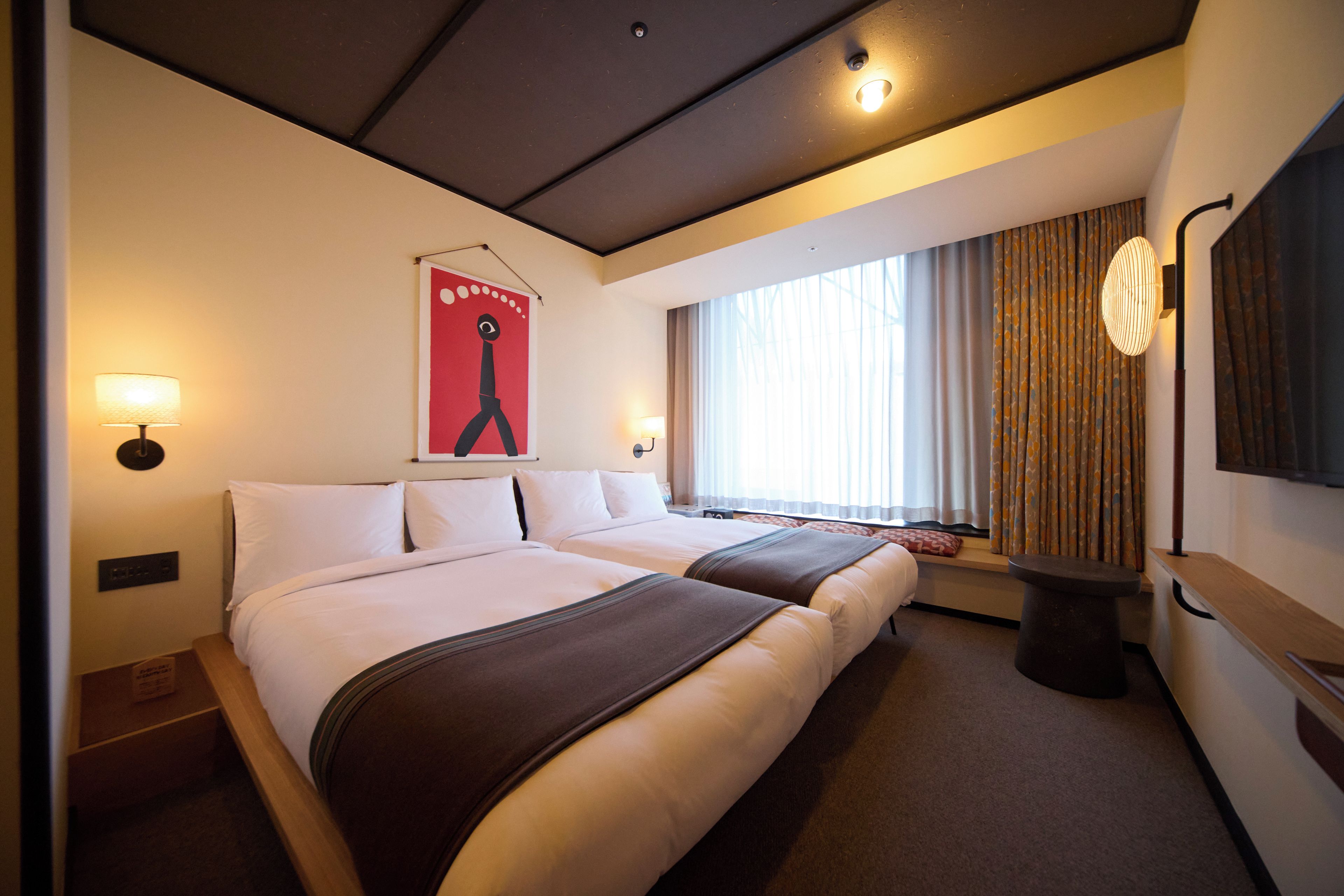 A hotel room with two beds, a large window with curtains, a wall-mounted TV, and a poster on the wall.