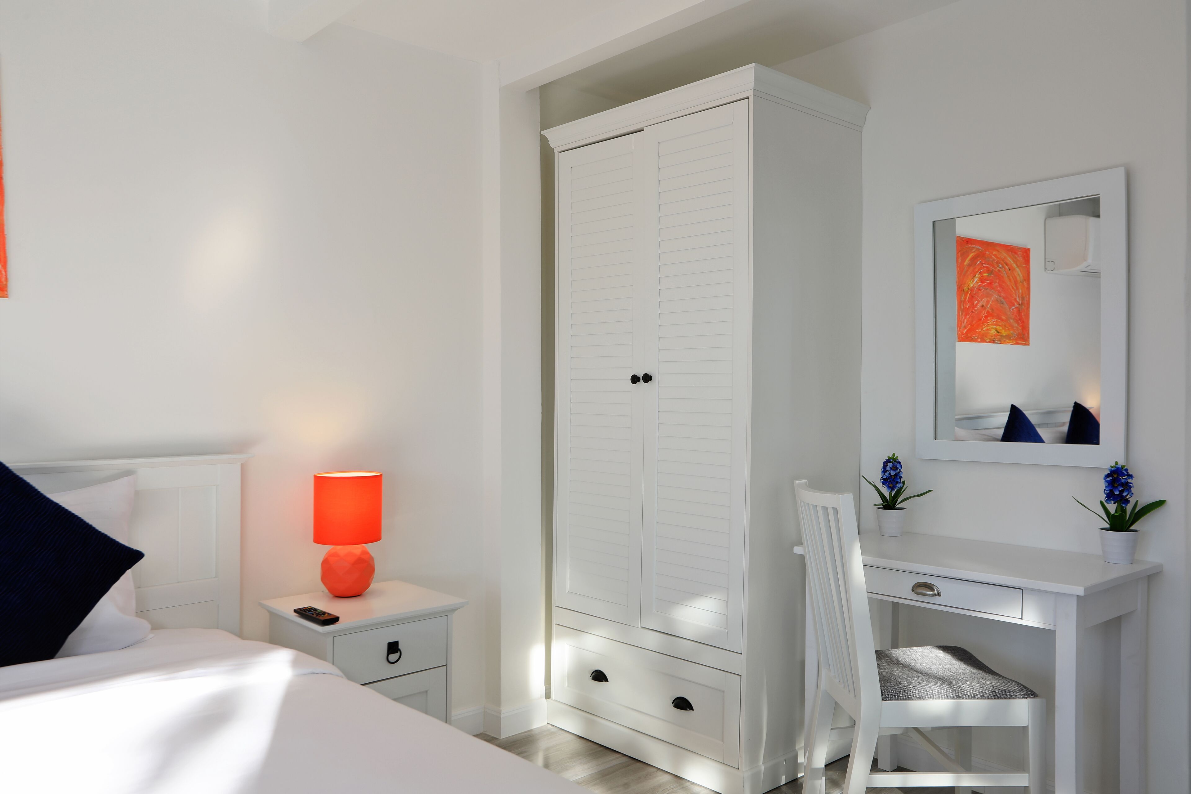 A bedroom with a bed, nightstand, lamp, wardrobe, desk, chair, and a mirror.