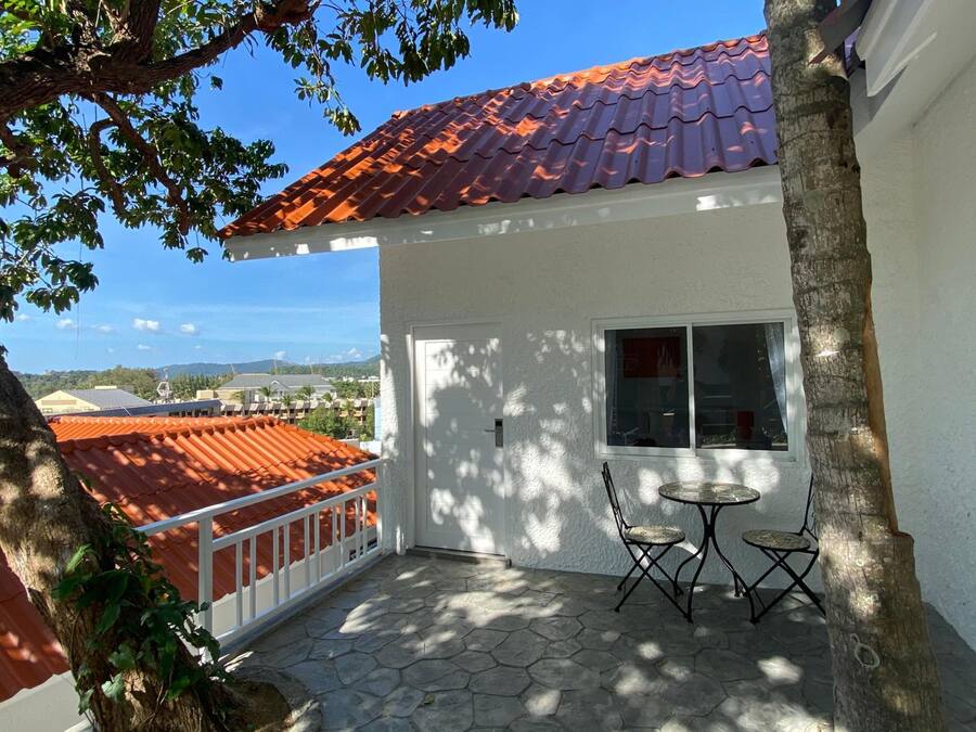 A small house with a red tiled roof, a white exterior, and a stone patio with two chairs.